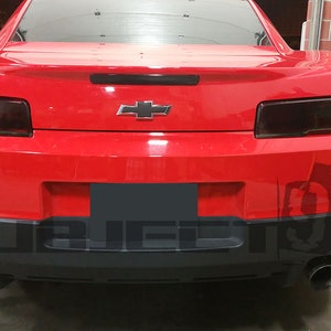 May include: Rear view of a red Chevrolet Camaro with tinted taillights and a black rear bumper. The car has a black license plate cover and dual exhaust tips.
