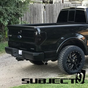 May include: Black pickup truck with a black bed and a black tailgate. The truck has a black rear bumper with a chrome hitch. The truck has black wheels with black and silver spokes. The truck is parked on a gravel driveway.