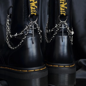 Op de afbeelding: Een paar zwarte enkellaarzen met dikke zolen en gele stiksels. Elke laars heeft een zwarte hiellus met gele AirWair-tekst, versierd met zilverkleurige metalen kettingen en een kleine zwaardbedel die aan een ronde ring hangt.