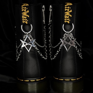 Peut inclure: Bottes en cuir noir avec coutures jaunes et l'inscription "AirWair" en jaune. Des breloques argentées avec des chaînes sont suspendues à l'arrière des bottes. Les breloques ont un motif en forme d'étoile.