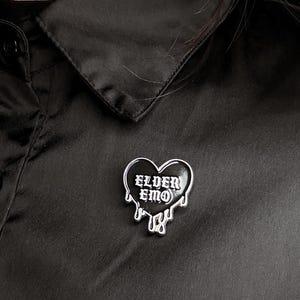 Può includere: Spilla nera a forma di cuore con le parole "ELDER EMO" in lettere gotiche bianche. La spilla ha un bordo argentato e un design a goccia. La spilla è attaccata a una camicia di raso nera.