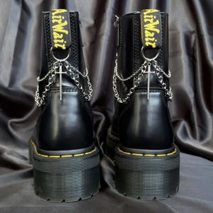 Puede incluir: Par de botas negras con plataforma, costuras amarillas y etiqueta amarilla "AirWair". Cada bota presenta una cadena plateada y un colgante de cruz. Las botas tienen una suela gruesa y están hechas de un material liso y brillante.