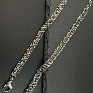 Peut inclure: Deux bracelets en cotte de mailles argentés avec fermoirs mousquetons. Les bracelets sont faits de petits anneaux métalliques imbriqués, créant une surface flexible et texturée. Les bijoux sont présentés sur une surface sombre et texturée.
