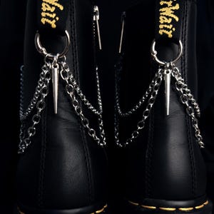 Puede incluir: Botas de cuero negro con cadenas y puntas plateadas. Las botas tienen una suela cosida amarilla y un lazo de tela negra y amarilla en la parte superior. Las cadenas cuelgan de un anillo plateado sujeto a la bota.
