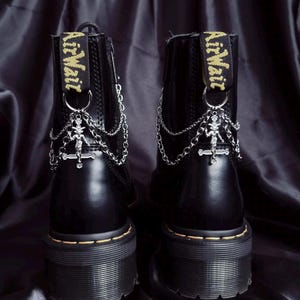 Puede incluir: Un par de botas de cuero negro con suelas gruesas y costuras amarillas. Cada bota presenta una trabilla negra en el talón con el texto amarillo AirWair y un accesorio de cadena de metal plateado con un dije en forma de cruz en la parte trasera.