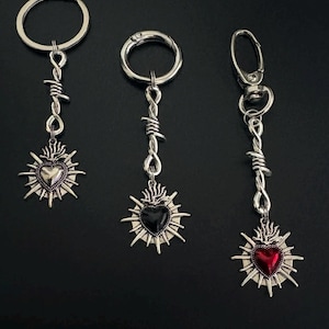 May include: Three silver keyrings with heart-shaped pendants. Each pendant features a different coloured heart: silver, black, and red. The pendants are surrounded by a sunburst design and attached to a barbed wire detail.