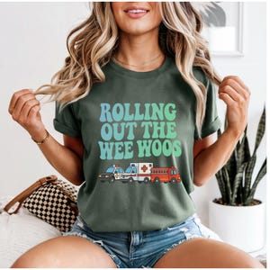 Puede incluir: Camiseta verde con el texto "Rolling Out The Wee Woos" en azul y verde. Debajo del texto hay ilustraciones de un coche de policía, una ambulancia y un camión de bomberos.