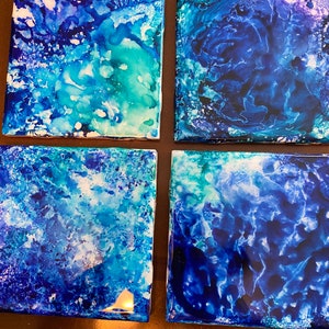 May include: Four square ceramic coasters with an abstract design in shades of blue and white. The coasters feature a swirling, marbled pattern in shades of blue, teal, and white.