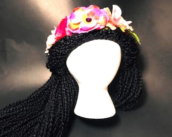 May include: A white mannequin head wearing a black crocheted headpiece with long black braided hair extensions. The headpiece is adorned with a crown of vibrant artificial flowers in shades of pink, orange, and white with green leaves.