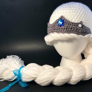 May include: White crocheted hat with a gray crocheted crown featuring a blue star-shaped gem. A long white braided yarn tail is attached to the hat.