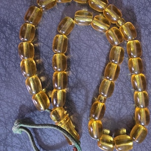 May include: A string of translucent, honey-coloured, barrel-shaped beads arranged in a loop. The beads are connected by a thin, green cord that is knotted at one end, featuring a small, decorative copper-coloured wire accent near the knot.