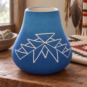 May include: A matte blue ceramic vase with a wide, rounded base and a tapered neck. The front of the vase features a white, hand-painted geometric design consisting of interconnected triangles and sharp, angular lines centered on the surface.