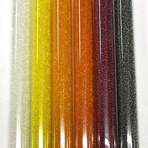 May include: Six small tubes of 11/0 size seed beads in various transparent colors, including clear, yellow, orange, red, and gray. Each tube contains 30 grams of beads and is labeled with a product number and price. The tubes are arranged in a row from left to right, with the clear beads on the left and the gray beads on the right.