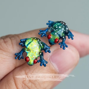 May include: Two small frog-shaped stud earrings featuring vibrant blue and green bodies with white spots and prominent red eyes. The frogs have detailed limbs and textured surfaces. The text Tiny Imaginarium is visible at the bottom centre.