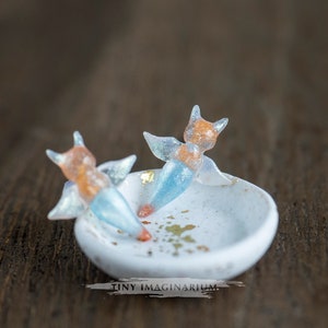 Pu&ograve; includere: Un piccolo piatto in ceramica bianca con due figurine a forma di volpe in miniatura. Le figurine hanno corpi arancioni e blu con ali traslucide. Dettagli in foglia d'oro sul piatto e sulla superficie in legno.