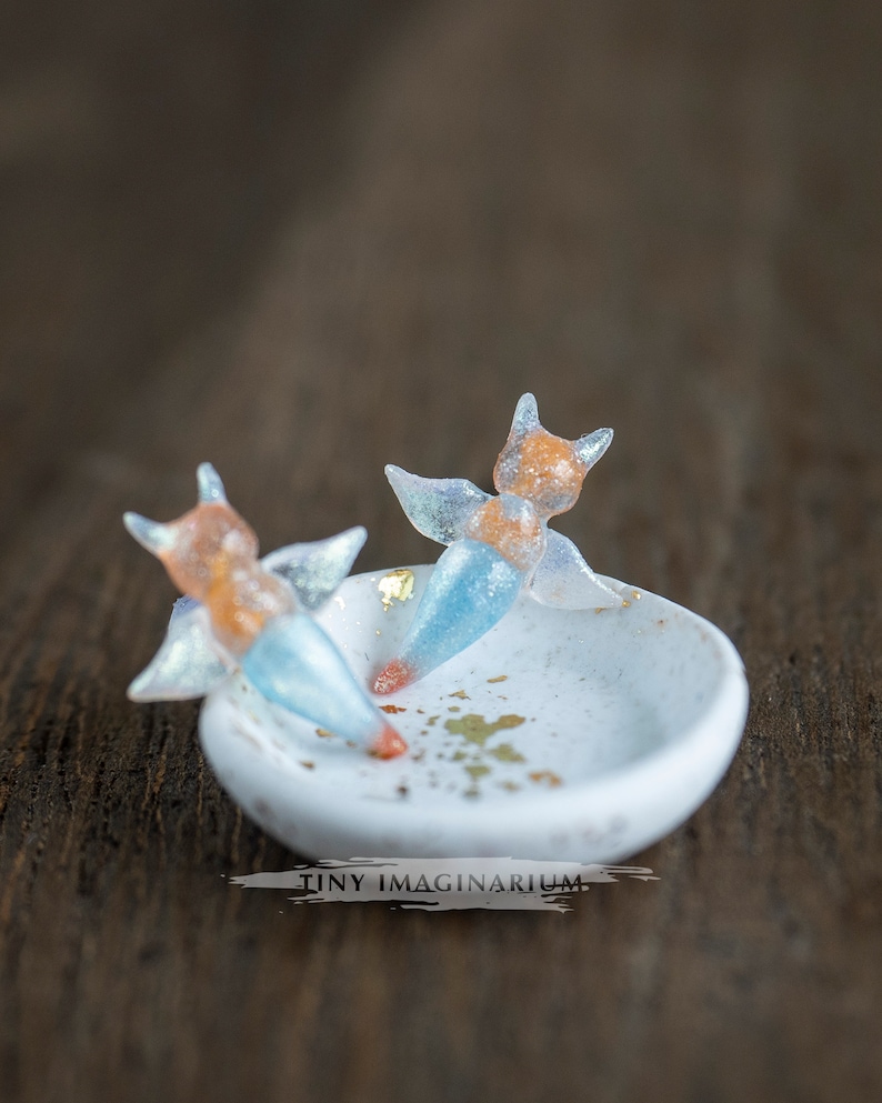 Pu&ograve; includere: Un piccolo piatto in ceramica bianca con due figurine a forma di volpe in miniatura. Le figurine hanno corpi arancioni e blu con ali traslucide. Dettagli in foglia d'oro sul piatto e sulla superficie in legno.