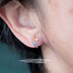 Pu&ograve; includere: Primo piano di un orecchino traslucido a forma di lumaca di mare. L'orecchino &egrave; arancione, blu e bianco e viene indossato su un orecchio. Il testo "TINY IMAGINARIUM" &egrave; sotto. Un gioiello unico.