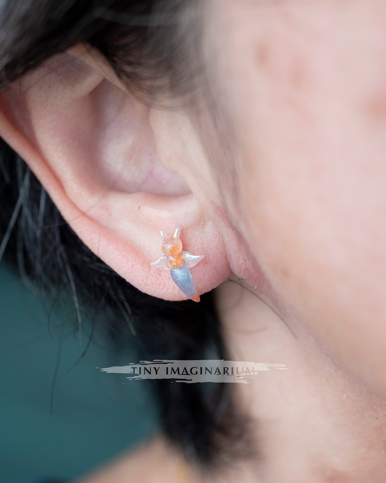 Pu&ograve; includere: Primo piano di un orecchino traslucido a forma di lumaca di mare. L'orecchino &egrave; arancione, blu e bianco e viene indossato su un orecchio. Il testo "TINY IMAGINARIUM" &egrave; sotto. Un gioiello unico.