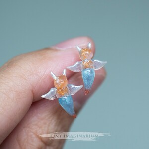 Pu&ograve; includere: Due figurine di angeli traslucidi in miniatura. Ogni figurina ha una testa arancione con piccole corna, ali trasparenti e un corpo blu. Le punte dei corpi sono rosse. Le figurine sono tenute in una mano.