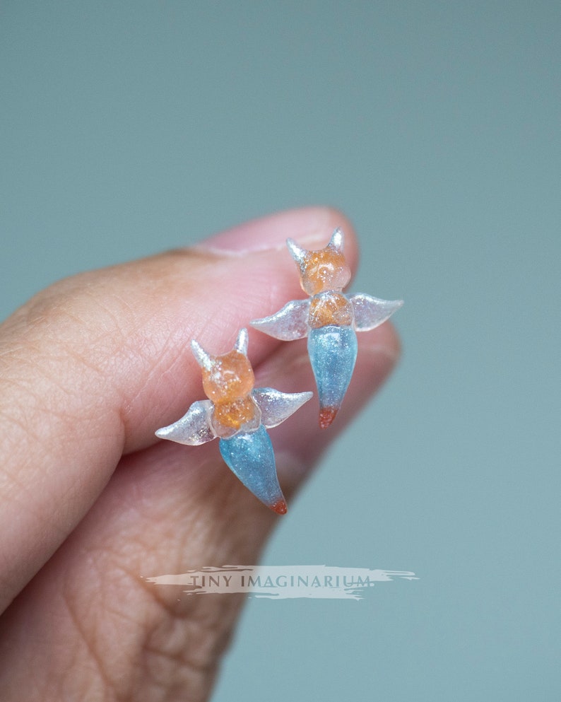 Pu&ograve; includere: Due figurine di angeli traslucidi in miniatura. Ogni figurina ha una testa arancione con piccole corna, ali trasparenti e un corpo blu. Le punte dei corpi sono rosse. Le figurine sono tenute in una mano.