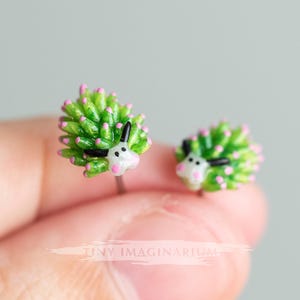 Puede incluir: Un par de pendientes de botón con forma de babosas marinas, con un cuerpo verde brillante con puntas rosadas, antenas negras y caras blancas. El texto TINY IMAGINARIUM es visible en el centro. Estos pequeños accesorios detallados tienen un acabado brillante y están diseñados para parecer nudibranquios.