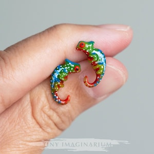 May include: A pair of colourful chameleon earrings, each with red, green, blue and white paint details. The earrings are small and delicate, and they are perfect for adding a touch of whimsy to any outfit.