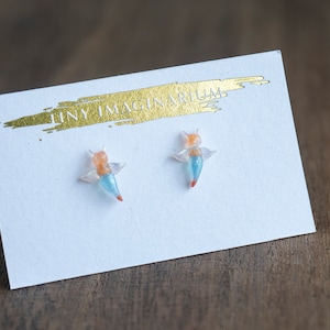 Pu&ograve; includere: Orecchini a forma di angelo di mare su una carta bianca con testo dorato "TINY IMAGINARIUM". Gli orecchini sono sculture in miniatura con ali arancioni, blu e trasparenti. Sono su una superficie di legno.