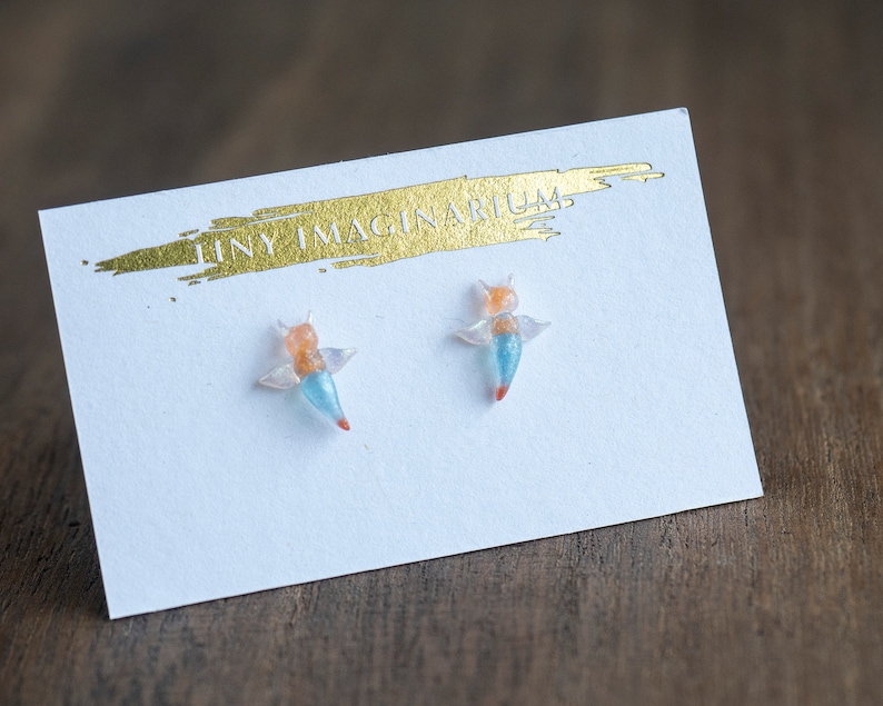 Pu&ograve; includere: Orecchini a forma di angelo di mare su una carta bianca con testo dorato "TINY IMAGINARIUM". Gli orecchini sono sculture in miniatura con ali arancioni, blu e trasparenti. Sono su una superficie di legno.