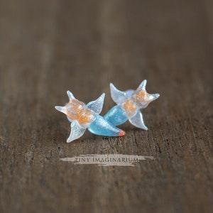 Pu&ograve; includere: Un paio di orecchini fatti a mano con un design a forma di stella. Gli orecchini sono traslucidi con dettagli arancioni e blu. Il testo "TINY IMAGINARIUM" &egrave; visibile. Sono su uno sfondo di legno.