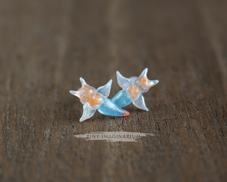 Pu&ograve; includere: Un paio di orecchini fatti a mano con un design a forma di stella. Gli orecchini sono traslucidi con dettagli arancioni e blu. Il testo "TINY IMAGINARIUM" &egrave; visibile. Sono su uno sfondo di legno.