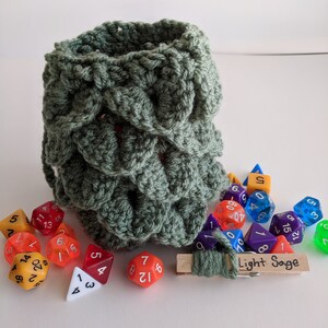 Op de afbeelding: Een groene gehaakte dobbelstenen tas met een houten wasknijper aan de bovenkant. De tas is gevuld met kleurrijke dobbelstenen, waaronder een rode d4, een rode d6, een gele d6, een blauwe d6, een paarse d6, een groene d6, een gele d8, een gele d10, een paarse d10, een gele d12, een paarse d12, een gele d20 en een groene d20. De wasknijper heeft een label met de tekst "Light Sage".