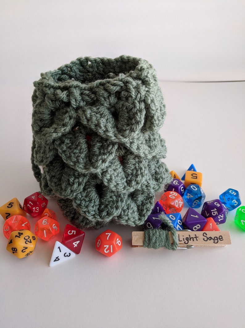 Op de afbeelding: Een groene gehaakte dobbelstenen tas met een houten wasknijper aan de bovenkant. De tas is gevuld met kleurrijke dobbelstenen, waaronder een rode d4, een rode d6, een gele d6, een blauwe d6, een paarse d6, een groene d6, een gele d8, een gele d10, een paarse d10, een gele d12, een paarse d12, een gele d20 en een groene d20. De wasknijper heeft een label met de tekst "Light Sage".