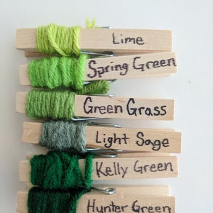 Op de afbeelding: Zes garenbollen in verschillende tinten groen, elk om een houten wasknijper gewikkeld. De wasknijpers zijn gelabeld met de namen van de groene tinten: Lime, Spring Green, Green Grass, Light Sage, Kelly Green en Hunter Green.