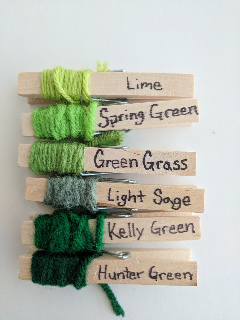 Op de afbeelding: Zes garenbollen in verschillende tinten groen, elk om een houten wasknijper gewikkeld. De wasknijpers zijn gelabeld met de namen van de groene tinten: Lime, Spring Green, Green Grass, Light Sage, Kelly Green en Hunter Green.