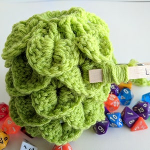 Op de afbeelding: Een limoengroene gehaakte dobbelstenen tas met een houten wasknijper met het label "Lime" eraan bevestigd. De tas is gevuld met kleurrijke dobbelstenen.