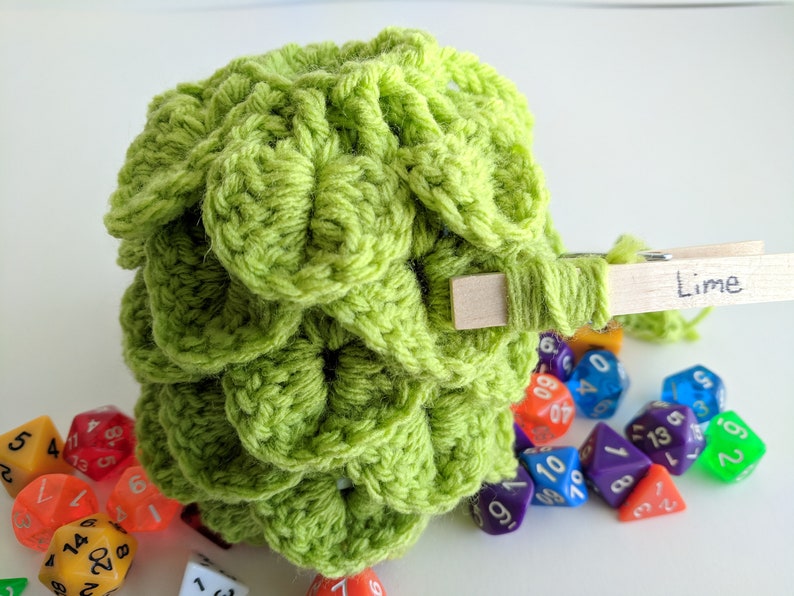 Op de afbeelding: Een limoengroene gehaakte dobbelstenen tas met een houten wasknijper met het label "Lime" eraan bevestigd. De tas is gevuld met kleurrijke dobbelstenen.