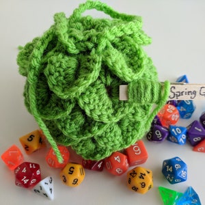 Op de afbeelding: Een groene gehaakte dobbelstenen tas met een houten label waarop "Spring Green" staat. De tas is gevuld met kleurrijke dobbelstenen.
