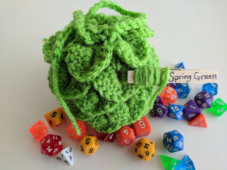 Op de afbeelding: Een groene gehaakte dobbelstenen tas met een houten label waarop "Spring Green" staat. De tas is gevuld met kleurrijke dobbelstenen.