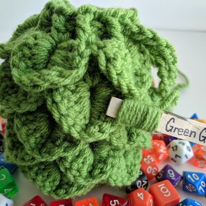 Op de afbeelding: Groene gehaakte dobbelsteen tas met een houten wasknijper met het label "Green Grass" en omringd door kleurrijke dobbelstenen.