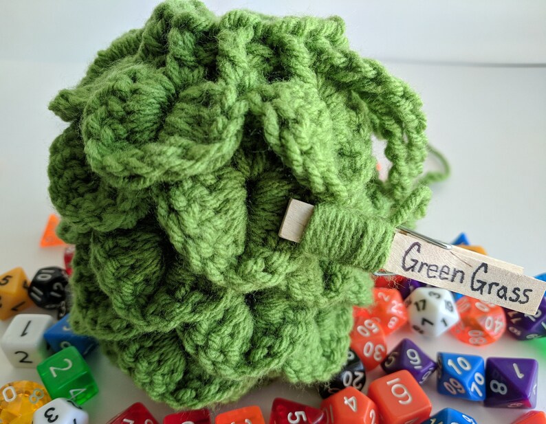 Op de afbeelding: Groene gehaakte dobbelsteen tas met een houten wasknijper met het label "Green Grass" en omringd door kleurrijke dobbelstenen.