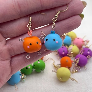 May include: A pair of dangle earrings featuring small, colorful clay figures with faces. The figures are attached to gold-colored hooks. The figures are in various colors, including orange, blue, green, yellow, and purple.