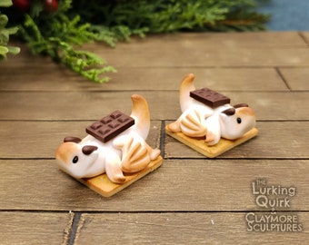 May include: Two whimsical clay sculptures of a creature resembling a squirrel, each lying on a square cracker. A dark chocolate bar rests on the creature's belly. The sculptures are white, brown, and tan, with black eyes. The background is a wooden surface.