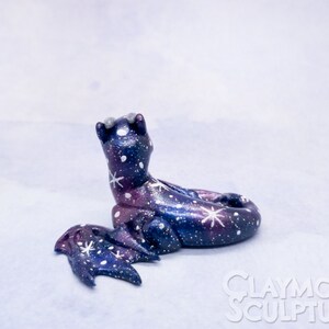 May include: A small, hand-painted ceramic dragon sculpture with a galaxy design. The dragon is curled up with its wings spread out. The sculpture is painted in shades of blue, purple, and white with white stars and swirls.