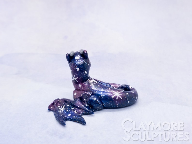 May include: A small, hand-painted ceramic dragon sculpture with a galaxy design. The dragon is curled up with its wings spread out. The sculpture is painted in shades of blue, purple, and white with white stars and swirls.