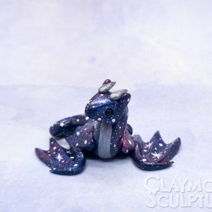 May include: A small, galaxy-themed dragon sculpture with a blue, purple, and silver paint scheme. The dragon has white stars painted on its body and wings. The sculpture is sitting on a light blue background.