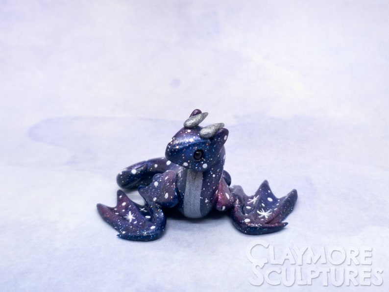 May include: A small, galaxy-themed dragon sculpture with a blue, purple, and silver paint scheme. The dragon has white stars painted on its body and wings. The sculpture is sitting on a light blue background.