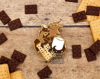 May include: A gold-tone enamel pin featuring a brown dragon holding a white marshmallow and a graham cracker. The dragon is sitting on a brown graham cracker. The pin says "Working & Quirk" and "Claymore Sculptures".