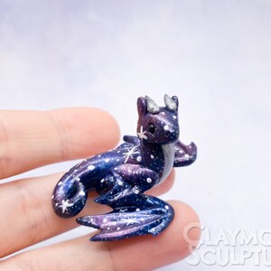 May include: A small, galaxy-themed dragon figurine with purple, blue, and white paint. The dragon has a curled tail, wings, and a star on its forehead.