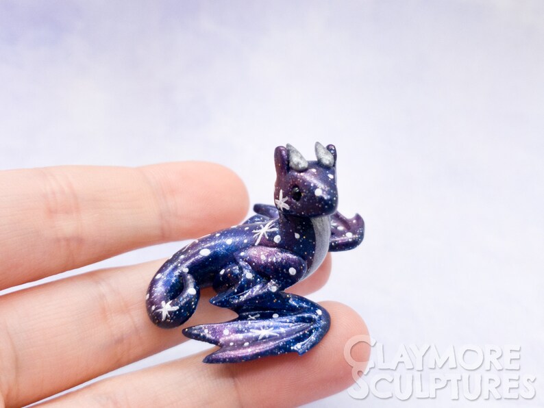 May include: A small, galaxy-themed dragon figurine with purple, blue, and white paint. The dragon has a curled tail, wings, and a star on its forehead.
