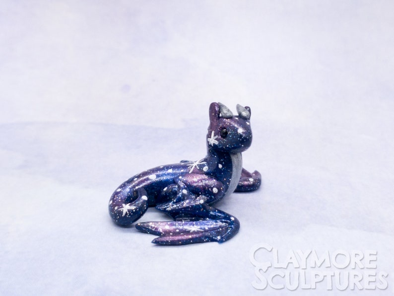 May include: A small, hand-crafted, galaxy-themed dragon figurine with a purple and blue iridescent finish. The dragon has white stars and a silver horn on its head.